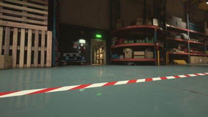 A dynamic view of a warehouse floor showcasing a strip of red and white caution tape. Shelves filled with boxes create a backdrop, indicating an organized yet busy environment during evening hours.