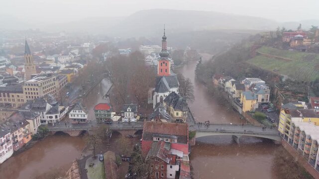 Cinematic drone reveal of the famous bridge houses in Bad Kreuznach during a winter storm. Flooded river and snowfall create a moody, textural atmosphere for travel and heritage themes.