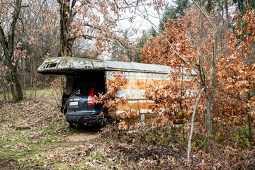 Improvised rural garage made from rusty bus body, sheltering a car in forest landscape during autumn winter.