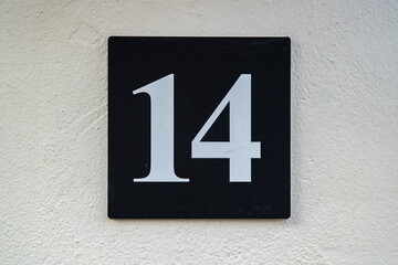 Close-up of metal house number fourteen sign mounted on white plaster wall, symbolizing urban address and navigation.