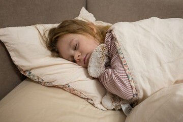 Peaceful little girl sleeping or resting with stuffed bunny toy in bed under white blanket at home