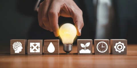 A hand gently places a glowing light bulb onto a line of wooden blocks displaying various symbolic icons representing innovation and progress
