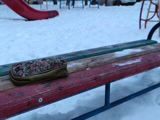 cosmetic bag on a wooden bench in a snowy playground. Concept of forgotten item, cold weather, winter season. Play area in winter.