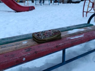 colorful cosmetic bag with floral pattern on a weathered wooden bench in a snow-covered playground during winter, depicting forgotten items.