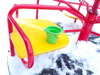 Green bucket on yellow merry go round with red handle covered with snow. Winter day at playground with frozen equipment. Outdoor play.