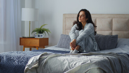 Caucasian pensive dreamy woman sitting on comfortable bed in cozy bedroom home thoughtful girl...