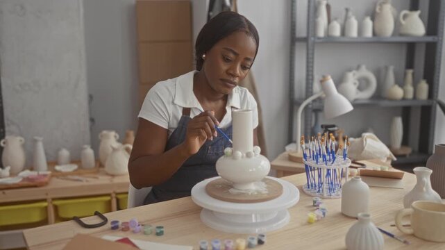 Woman potter painting a white ceramic vase with a fine brush in a pottery studio, hand steady as she applies glaze beads along the rim at a worktable; concentration craft.