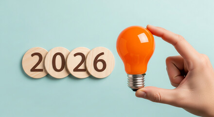 Image shows round wooden blocks with "2026" and a hand holding an orange bulb. Represents future, planning, and creative business idea for upcoming year