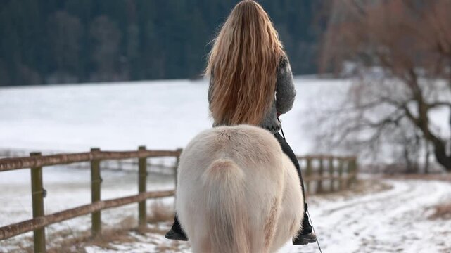 Young woman riding a white Icelandic horse along a snowy rural path, rear view showing calm winter countryside