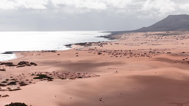 Aerial dunes, Atlantic surf, lava rock, and flat topped massif in Fuerteventura, Canary Islands. Narrow coastal road, tiny cars and beachgoers show scale under sunlit clouds.
