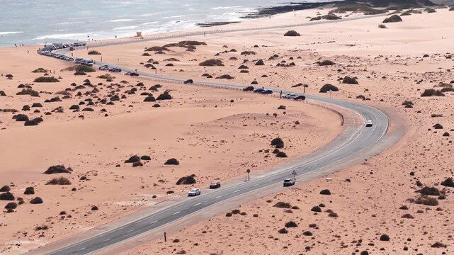 Aerial 4K scene shows a sinuous coastal road through orange dunes on Fuerteventura, Canary Islands, with parked cars, moving vehicles, surf, scrub, and tiny figures