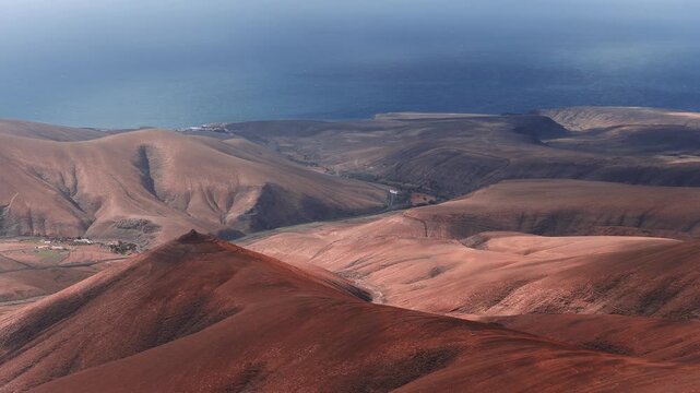 Aerial 4K glide shows rust red hills, a conical crater, dunes, and the Atlantic on Fuerteventura, Canary Islands, with a road, buildings, and Lanzarote on horizon.