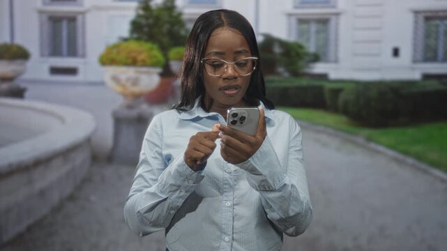 Woman tapping smartphone screen with finger, wearing glasses and a light shirt, standing by a building fountain and paved path; quiet concentration.