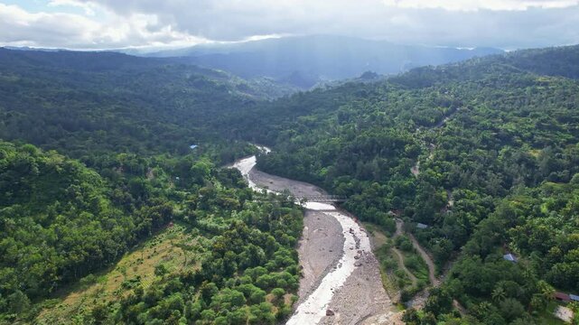 Aerial drone footage of a main wild river, with low waters, in the middle of Timor island, Indonesia, with mountains, sun rays, and everything covered by green lush natural forests