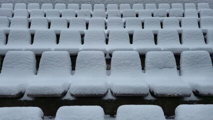 Rows of stadium seating completely covered in a thick, uniform layer of fresh white snow, creating a repetitive geometric pattern of soft, rounded shapes against a dim background.