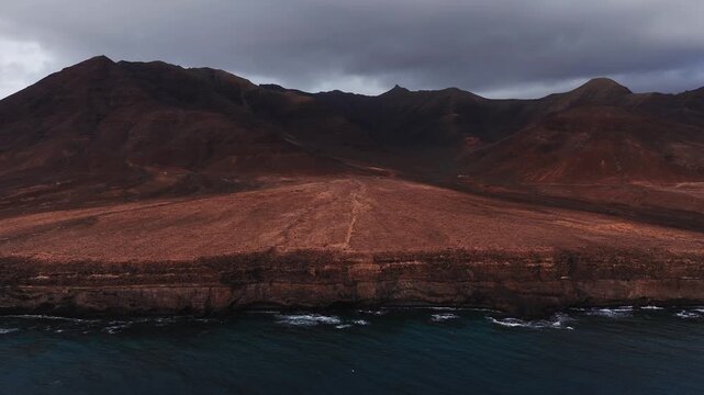 Aerial view of Fuerteventura shows rust coloured plain, steep layered sea cliffs, dark volcanic ridge with triangular peak, faint trail, surf, and overcast light.