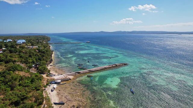 Aerial drone footage of Bolok ferry harbour area, in Kupang city, Timor island, Indonesia, with a small rocky jetty, clear turquoise and blue waters, a forest, and another island in the front