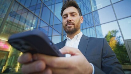 Bearded Caucasian male in elegant suit standing near business center entering message on smartphone screen with focused expression. Confident professional using technology outdoors on bright day.