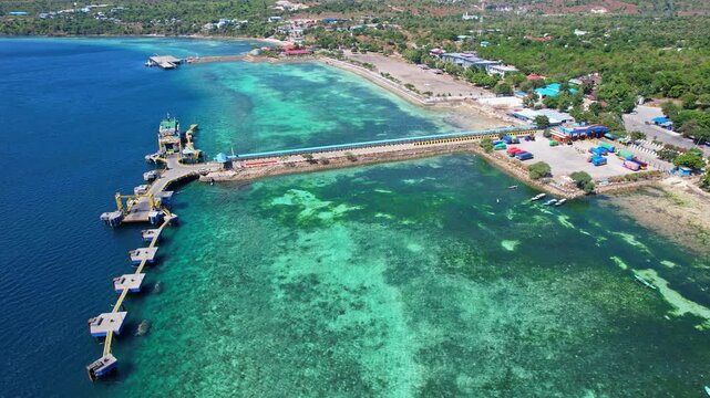Aerial drone footage of Bolok ferry harbour area, in Kupang city, Timor island, Indonesia, with a jetty, clear turquoise and blue waters, corals and reef, some buildings, a parking and a ferry boat