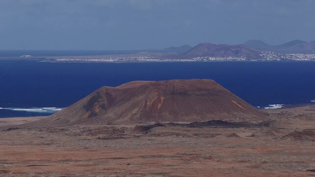 Aerial drone pan over a flat topped volcanic cone in Fuerteventura, rust and black lava textures, Atlantic shore, coastal town, Lanzarote on the horizon in daylight