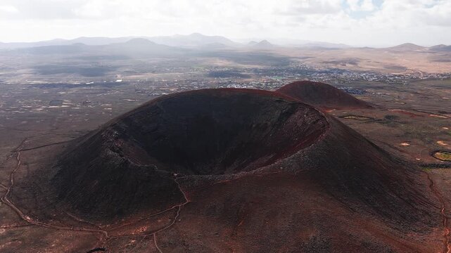 Aerial 4K sweep passes a sharp volcanic crater, red black cinder cones, lava fields, paths, stone enclosures, and a distant town in Fuerteventura, Canary Islands.