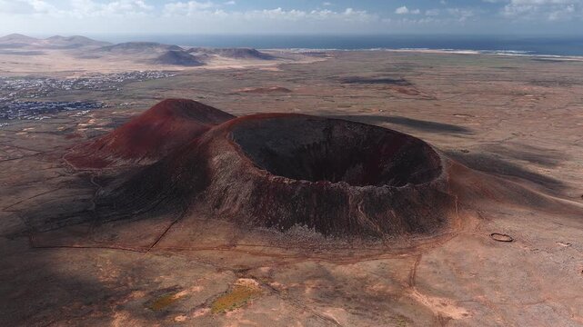Aerial view shows a bowl shaped volcanic crater in Fuerteventura, red and black scoria slopes, a secondary cone, stone walls, enclosures, a coastal village, and the Atlantic.