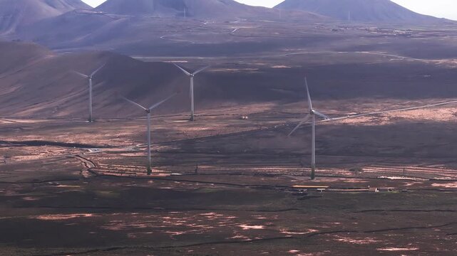 Aerial view shows wind turbines on lava plains and cones in Fuerteventura, Canary Islands. Camera glides and pans as blades spin, roads and highway trace the terrain.