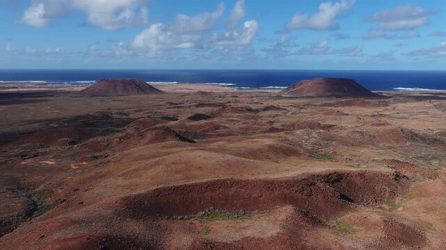 Aerial 4K drone glide over rust red basaltic plain on Fuerteventura, Canary Islands, revealing small craters, two flat topped cones, surf, and midday clouds.