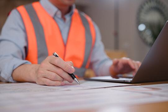 Engineer wearing safety vest writing on documents while using laptop at work desk. Concept of construction planning, project management, engineering analysis, safety, and professional workflow.