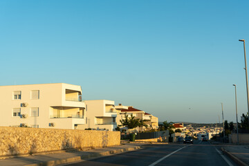 Modern white apartment buildings with balconies and air conditioning along road in Mediterranean coastal town at golden hour. Concept of resort real estate, new development, vacation property, travel.