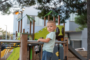 Obraz premium Blonde boy in striped shirt standing on playground platform holding railing and looking at camera in resort park with colorful structures. Concept of childhood portrait, summer vacation, outdoor fun.