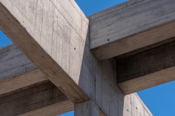 Massive concrete beams intersect beneath a clear blue sky, revealing stark geometric angles and texture across