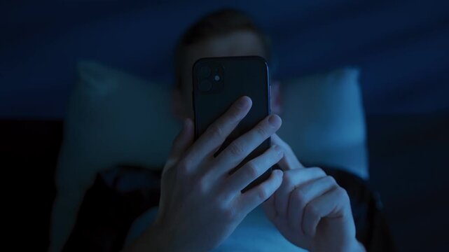Close up of man with a smartphone in his hands, illuminated with blue light of the screen, browsing social media and consuming content late at night instead of sleeping. Digital addiction and insomnia