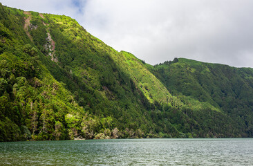 Fototapeta premium Lush green mountains surrounding Sete Cidades lagoon in Azores, Portugal. Crater lake with vibrant greenery and clear blue waters. Volcanic landscape with scenic views of Sao Miguel island.