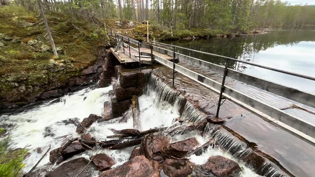 water cascades stone weir into rocky stream below wooden footbridge fallen logs moss covered cliffs surround calm norway nature forest cascading boreal woodland 