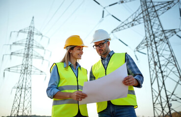 Two engineers in safety vests and helmets discuss project plans near power lines. They examine blueprints outdoor on sunny day at construction site. Focus on teamwork and engineering.