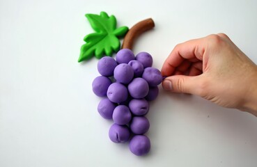 Hand crafts plasticine grapes on white surface. Fine motor skill learning activity in childcare center. Arts and crafts project for kids shows how to model fruit shapes with playdough.