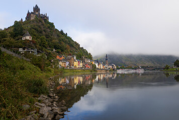 Fototapeta premium Reichsburg Cochem über der Mosel bei ruhigem Wasser, historische Altstadt von Cochem am Flussufer mit klarer Spiegelung, bewaldeter Hügel und dezenter Dunst in der Landschaft