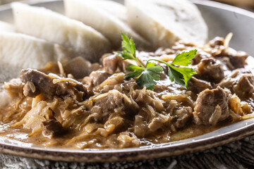 Traditional Szeged goulash with pieces of meat and sauerkraut is served on a plate, complemented by a few slices of light steamed dumplings and garnished with parsley