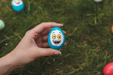 Hand holding a painted blue easter egg with a smiling face on green grass with colorful eggs with holiday with fun