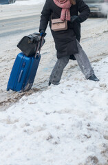 A young woman with a suitcase and bags crosses a snowy road. Winter in the city. Selective focus