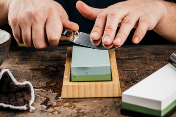 A man sharpens a knife with a grindstone on a rustic wooden table.