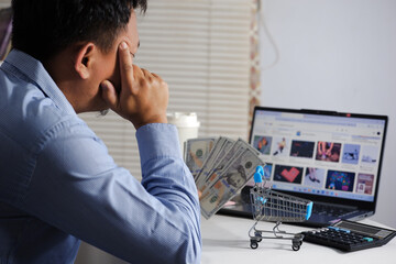 A person holds US dollar bills in front of a laptop while browsing an e-commerce payment screen, while holding their forehead, digital shopping themes, and money management visuals. Financial consider