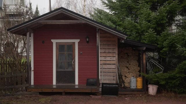 Red wooden storage shed with firewood and gas tanks during steady rain in Finland. Practical outbuilding designed for typical Scandinavian autumn weather conditions in Kotka region.