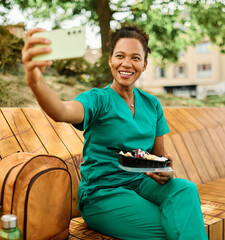 Young woman or a nurse or a doctor during office hours break enjoying a fresh salad and using mobile phone taking a selfie   in a park during a sunny day while smiling and relaxing on a bench