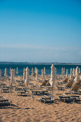 Visitors relax on sunbeds with umbrellas at a beach in Bulgaria. The coastline of sunny beach stretches along the water, while buildings rise in the distance. It is daytime and the sky is clear