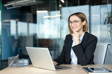 Fototapeta premium Young businesswoman wearing eyeglasses looking away and smiling while sitting at her modern office desk with a laptop. Embodying innovation. Success. And professional growth in a corporate environment