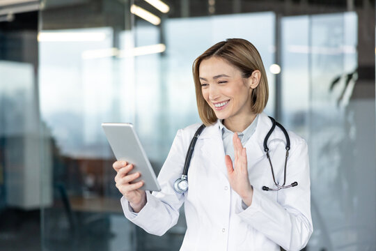Female doctor in lab coat and stethoscope smiling, waving hand while making a video call on a digital tablet, offering remote medical advice and online healthcare - Powered by Adobe