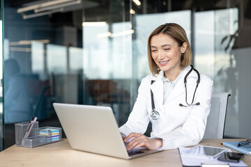 Young female doctor in a white coat and stethoscope smiling while typing on a laptop, providing an online consultation and telehealth services from a modern clinic office
