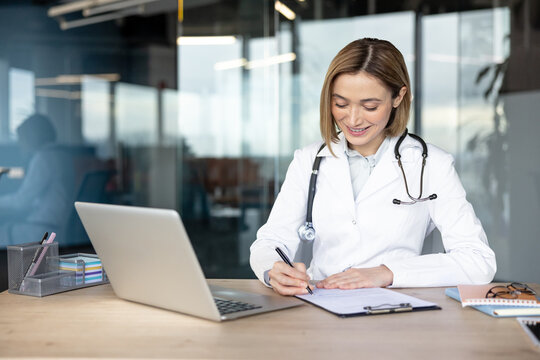 Female doctor in a white lab coat with a stethoscope around her neck smiling, sitting at an office desk, and writing a medical report or prescription on a clipboard with a laptop nearby